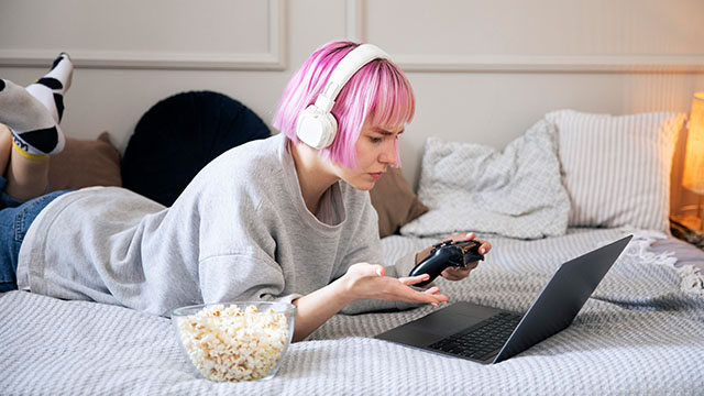 Devojka roze kose leži u spavaćoj sobi i igra igrice na laptopu koristeći džojstik