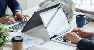 Dva biznismena sede u kancelariji za radnim stolom i koriste laptopove