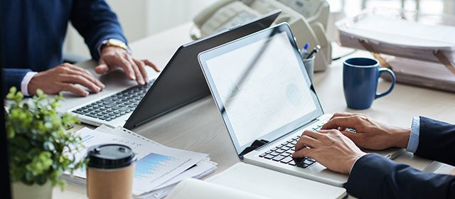 Dva biznismena sede u kancelariji za radnim stolom i koriste laptopove