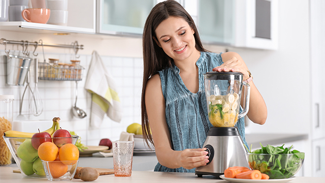 Nasmejana žena u kuhinji priprema zdrav obrok od voća i povrća u blenderu