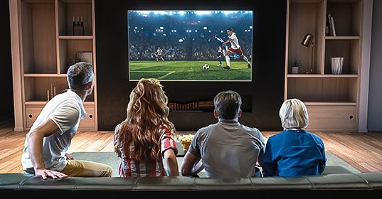 Grupa ljudi sedi ispred televizora i gleda fudbalsku utakmicu na velikom ekranu