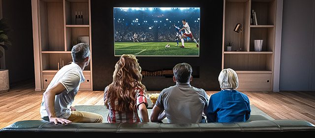 Grupa ljudi sedi ispred televizora i gleda fudbalsku utakmicu na velikom ekranu