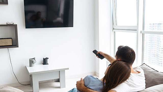 Zagrljen mladi par sedi na udobnom kauču i uključuje televizor postavljan na zidu ispred njih