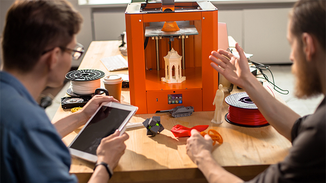 Dva čoveka istražuju i posmatraju proces 3D štampe ispred štampača u kome je odštampana figura