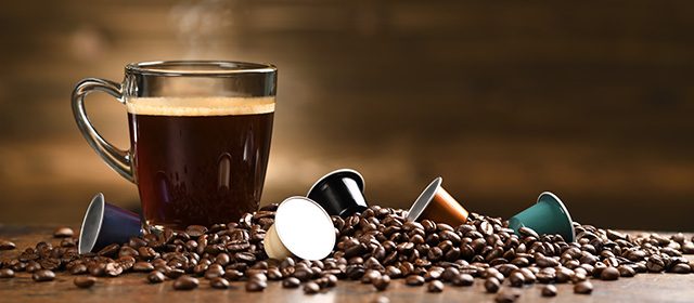 Na stolu se nalazi šoljica kafe, prosuta kafa u zrnu i nekoliko kapsula za kafu
