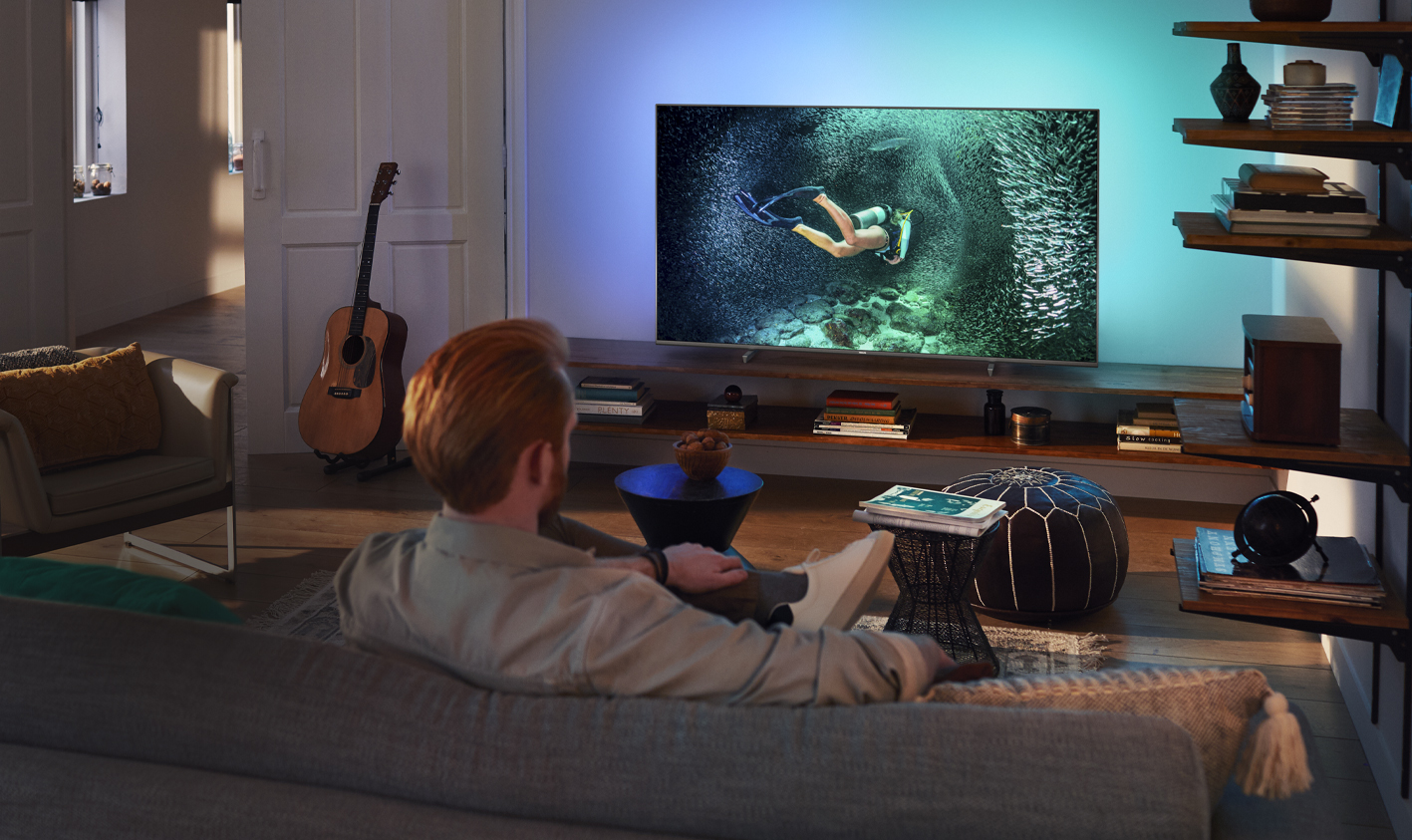 Muškarac slikan sa leđa sedi u dnevnoj sobi i gleda TV sa Ambilight osvetljenjem