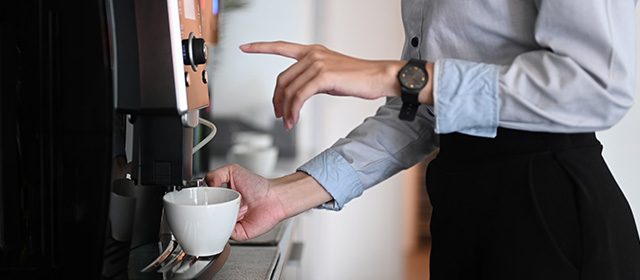 Devojka u elegantnoj košulji i suknji na poslu priprema kafu koristeći aparat za espresso