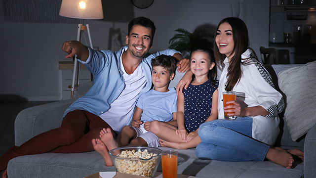 Mladi bračni par sa decom sedi na kauču, jede kokice i uživa u gledanju filma koristeći projektor